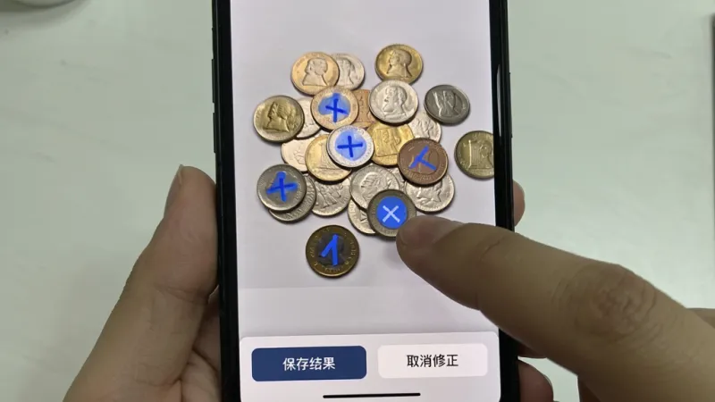 拍照扫描计数软件手动修正界面，手机屏幕显示一堆散落的硬币，部分硬币有蓝色标记，部分没有标记，用户手指正在点击一个未标记的硬币进行添加，界面下方有“保存结果”和“取消修正”按钮，浅灰色背景，风格简约，画质清晰