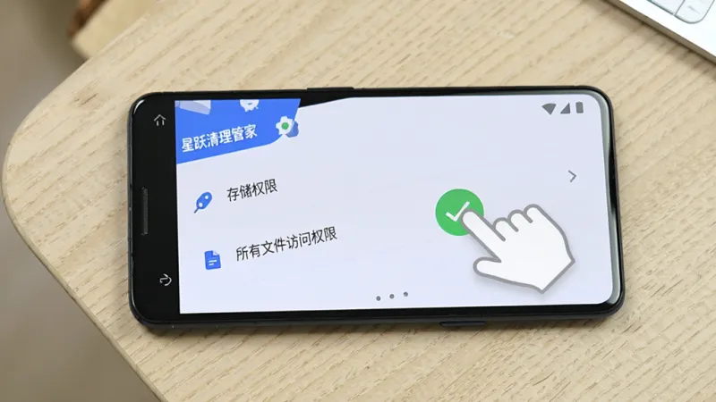 一部安卓手机放在桌面上,屏幕显示星跃清理管家的权限设置页面,页面中有“存储权限”和“所有文件访问权限”两个选项,旁边有手指点击允许的示意动画,背景是浅色木纹桌面,风格写实清晰,画质干净,文字标注“星跃清理管家