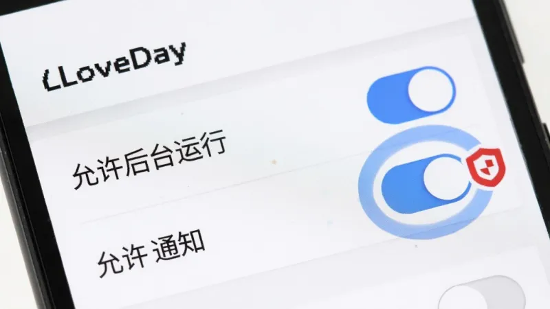 一部安卓手机屏幕特写,屏幕上显示“LoveDay”的设置页面,页面中有多个开关按钮,其中“允许后台运行”和“允许通知”的开关被高亮标注为蓝色,旁边有一个红色盾牌图标表示安全提示,界面干净整洁,背景是纯白色,画质清晰,无多余元素