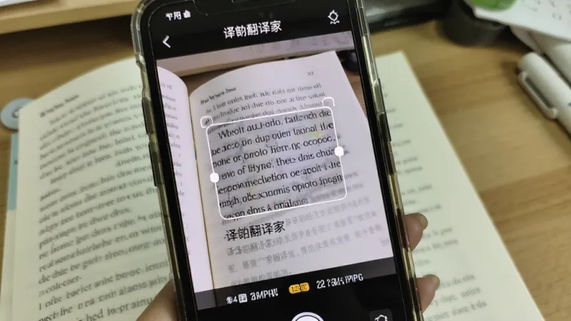 一张展示手机摄像头对着英文书页拍照的画面,手机屏幕上显示着“译劭翻译家”的拍照翻译界面,英文文字被识别后下方直接显示出对应的中文翻译,画面中有聚焦框,背景是书桌,风格真实自然