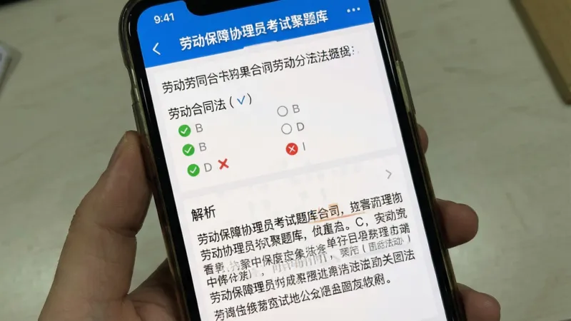 一张手机屏幕截图展示图,画面中手机屏幕上显示着“劳动保障协理员考试聚题库”的章节练习界面,屏幕上半部分是一道关于劳动合同法的单选题,选项下方有绿色对勾和红色叉号标记,屏幕下半部分是详细的文字解析区域,解析中标注了关键法条,整体界面以白色为底,蓝色作为点缀,文字清晰,排版整齐,画质干净,无多余元素