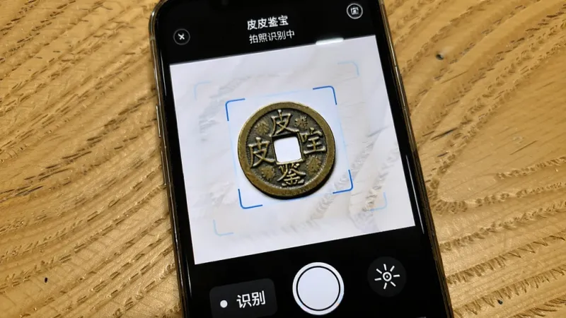 皮皮鉴宝拍照识别功能展示,主体是手机屏幕显示拍摄界面,布局为上方是取景框,中间有一个古钱币图案正在被识别,下方有识别按钮和闪光灯图标,排版清晰,风格为写实UI风格,细节上界面文字为皮皮鉴宝和拍照识别中,背景是木质桌面,画质干净,无多余元素