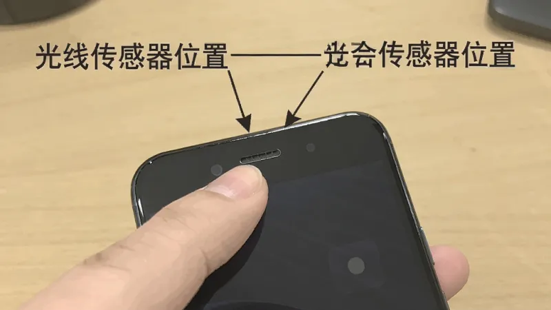一张手机顶部传感器位置的示意图,画面中一只手拿着手机,手指指向屏幕顶部听筒附近,旁边有箭头标注“光线传感器位置”,背景是纯色桌面,风格说明性,画质清晰,文字标注明确,无多余元素
