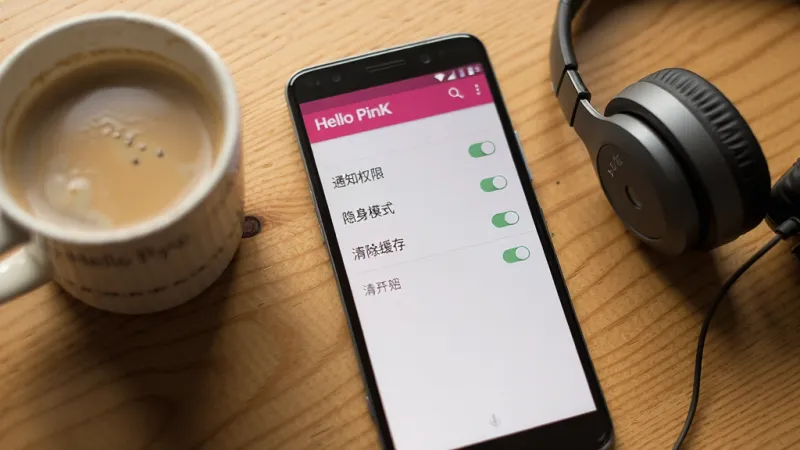 一部安卓手机屏幕上显示Hello PinK应用设置界面的截图,界面中包含“通知权限”“隐身模式”“清除缓存”等选项开关,旁边放着一杯咖啡和一副耳机,背景是木质桌面,风格生活化,光线柔和,画质干净清晰,无多余杂乱元素
